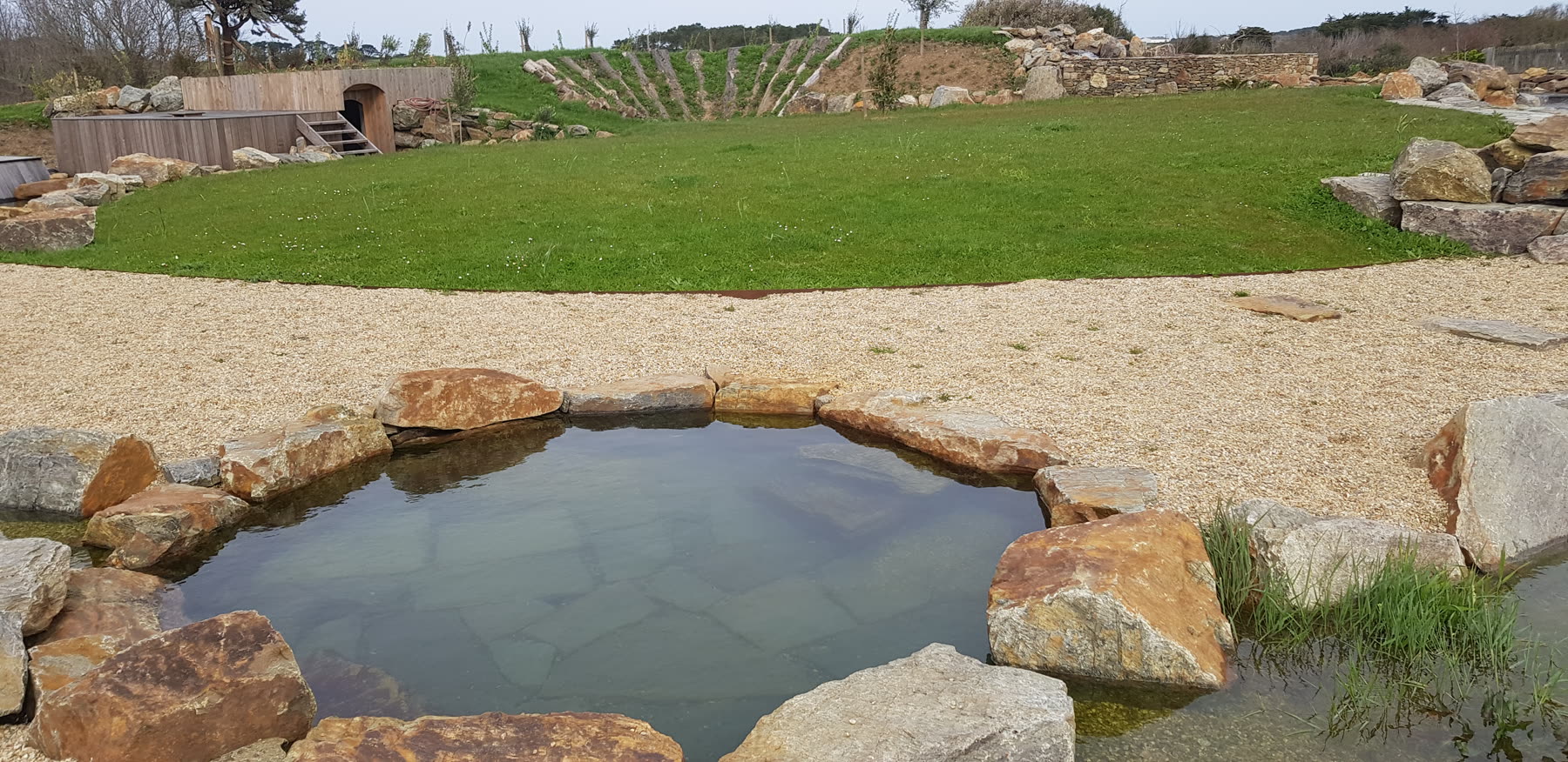 Panorama: le bassin de rochers au premier plan, la pelouse, une cabane en bois et des plantations en gradins au fond