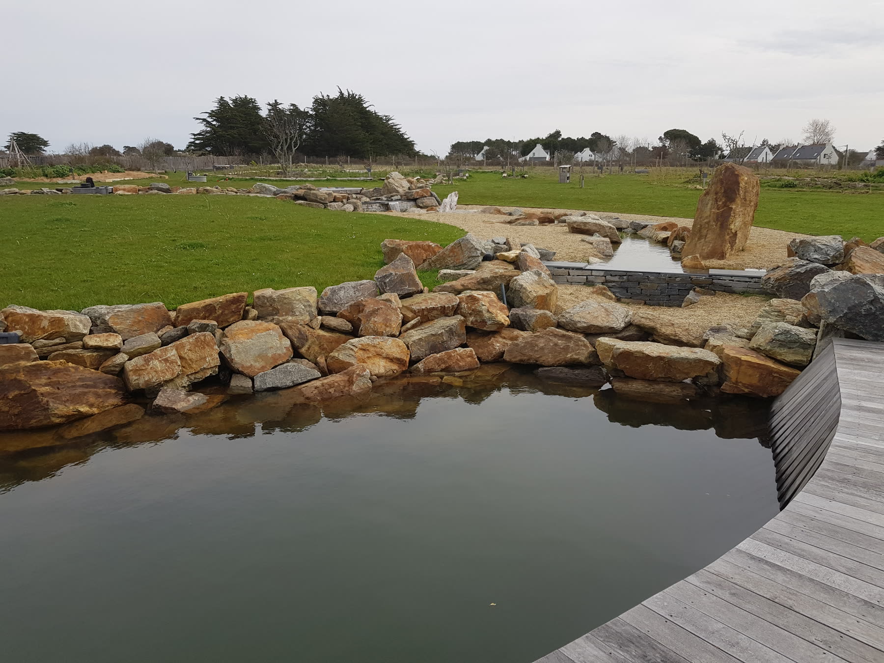 Le bassin de rochers et sa terrasse bois en courbe, la pelouse, les maisons blanches et les cyprès au fond