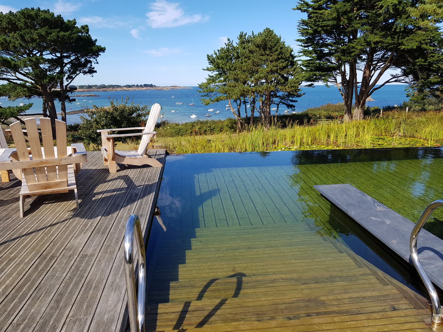 Deux fauteuils en bois sur la terrasse au bord de l’eau, échelle inox, la mer derrière les arbres