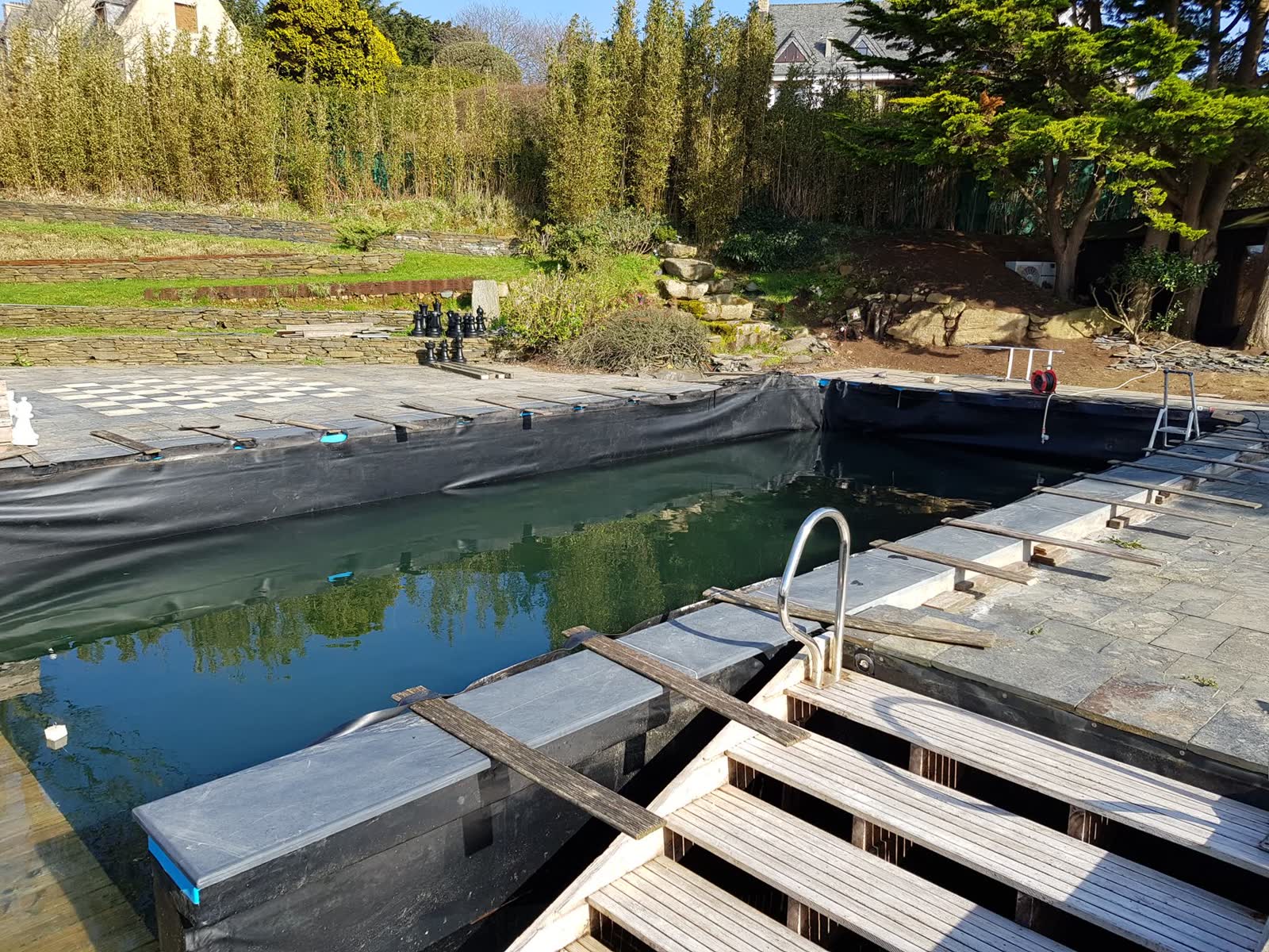 Le bassin en chantier vu depuis le ponton: membrane noire, planches de la terrasse en cours de pose, échelle déjà en place
