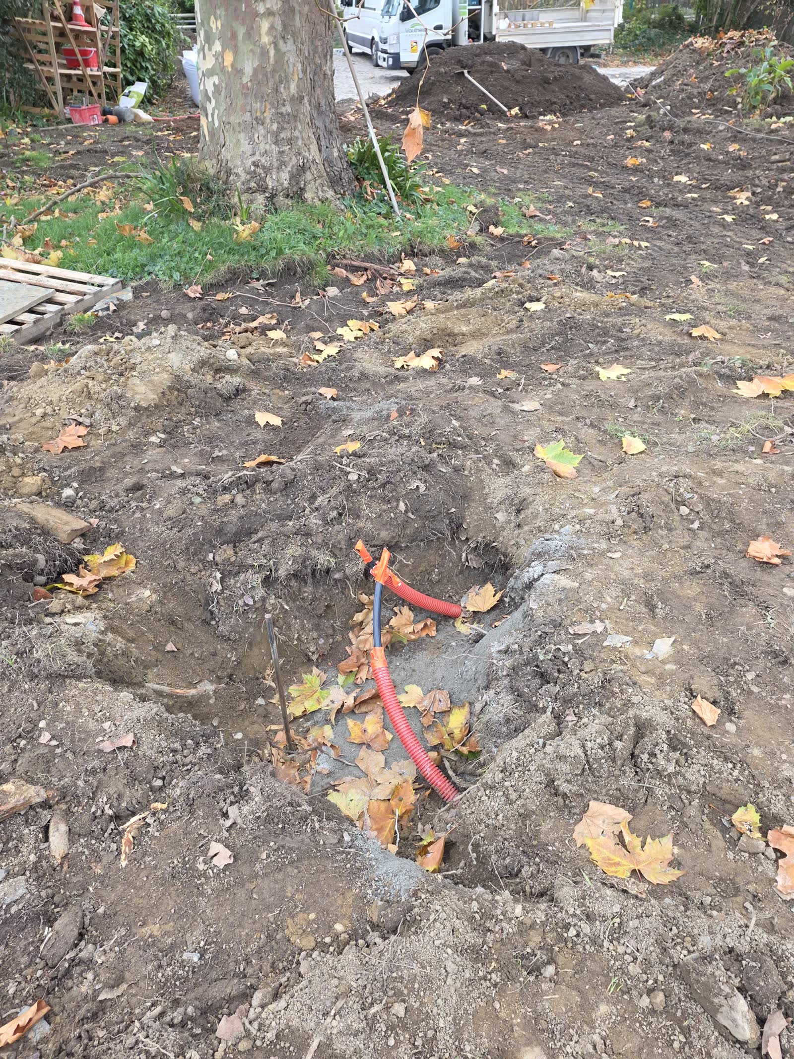 Une tranchée dans la terre couverte de feuilles mortes, une gaine électrique rouge et une conduite noire qui en sortent, un platane et un camion plateau blanc au fond
