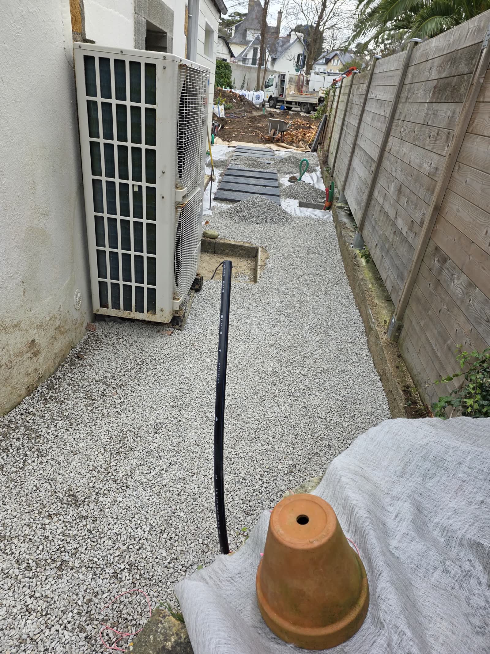 Une allée gravillonnée le long de la maison, une pompe à chaleur extérieure à gauche, une conduite noire posée sur le gravier, une clôture en bois à droite