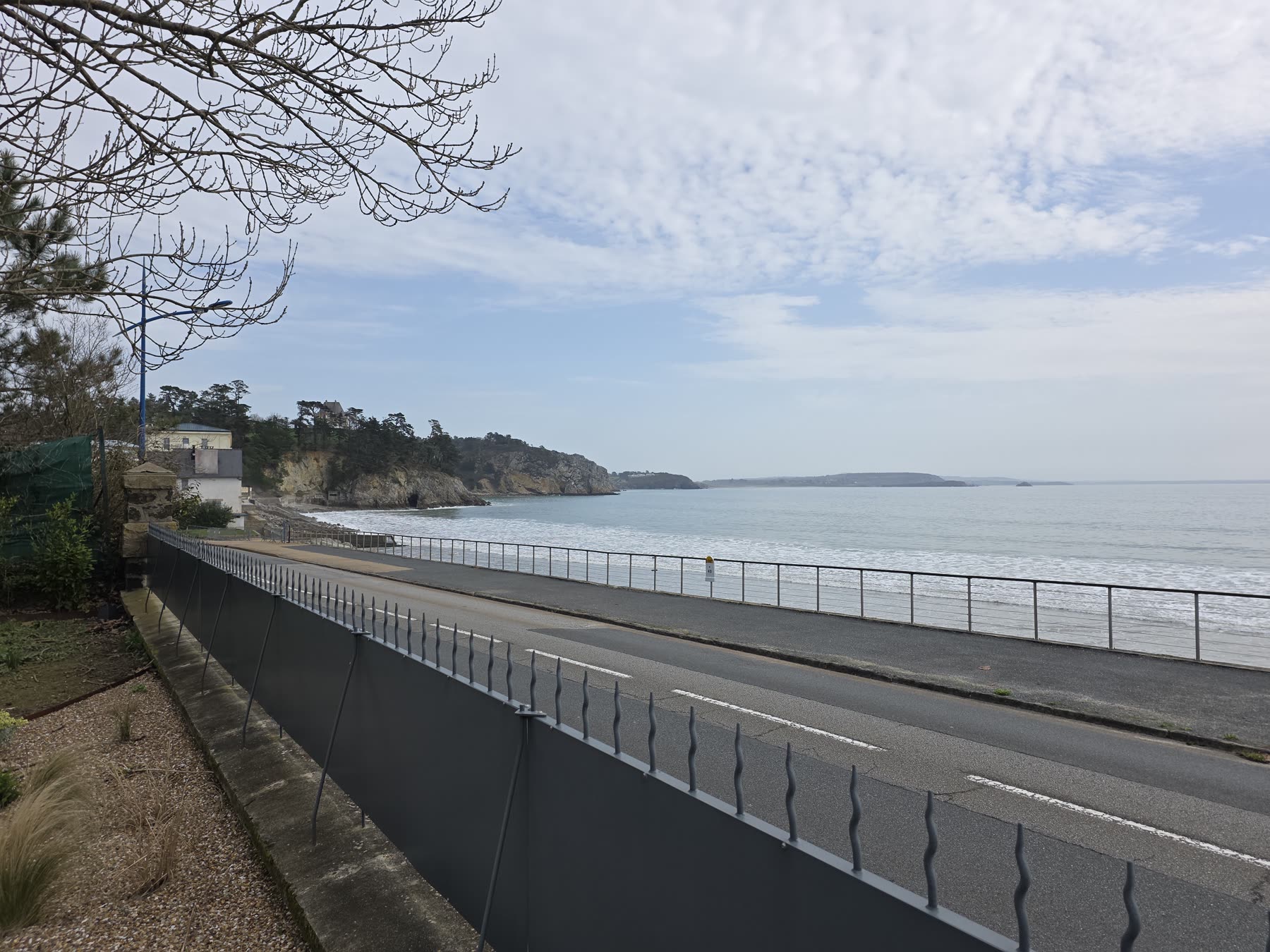 Depuis le jardin: un muret et une clôture grise le long de la route du front de mer, le garde-corps de la promenade, la plage, la baie et les falaises boisées au fond