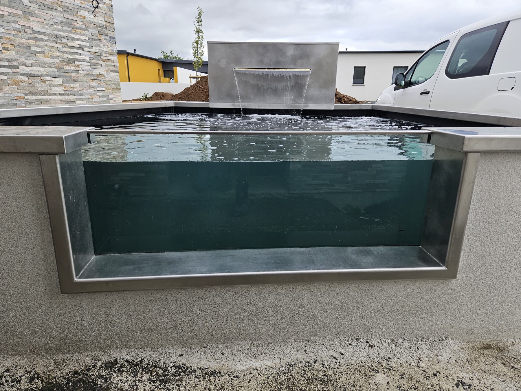 Le hublot vitré dans la paroi du bassin: on voit l’eau verte et la lumière à travers, la margelle inox au-dessus, l’enduit blanc autour