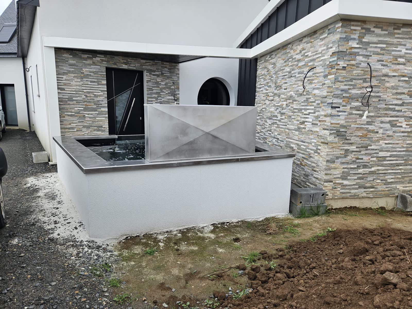 Le bassin vu de face depuis l’allée: le mur enduit, la margelle inox, la façade en pierre et le bardage noir de la maison