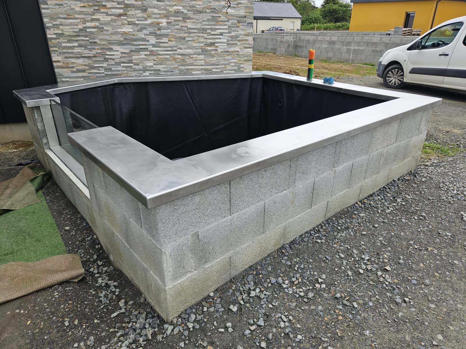 Le bassin en construction: parpaings, margelle inox posée, la réservation du hublot, gravier et terre autour