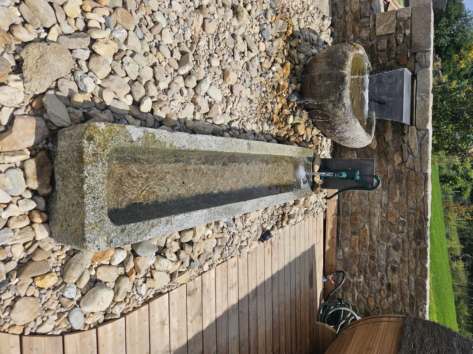 Une auge longue en granit et son bec inox, l’eau qui s’écoule, terrasse bois à droite, pelouse et maisons au fond
