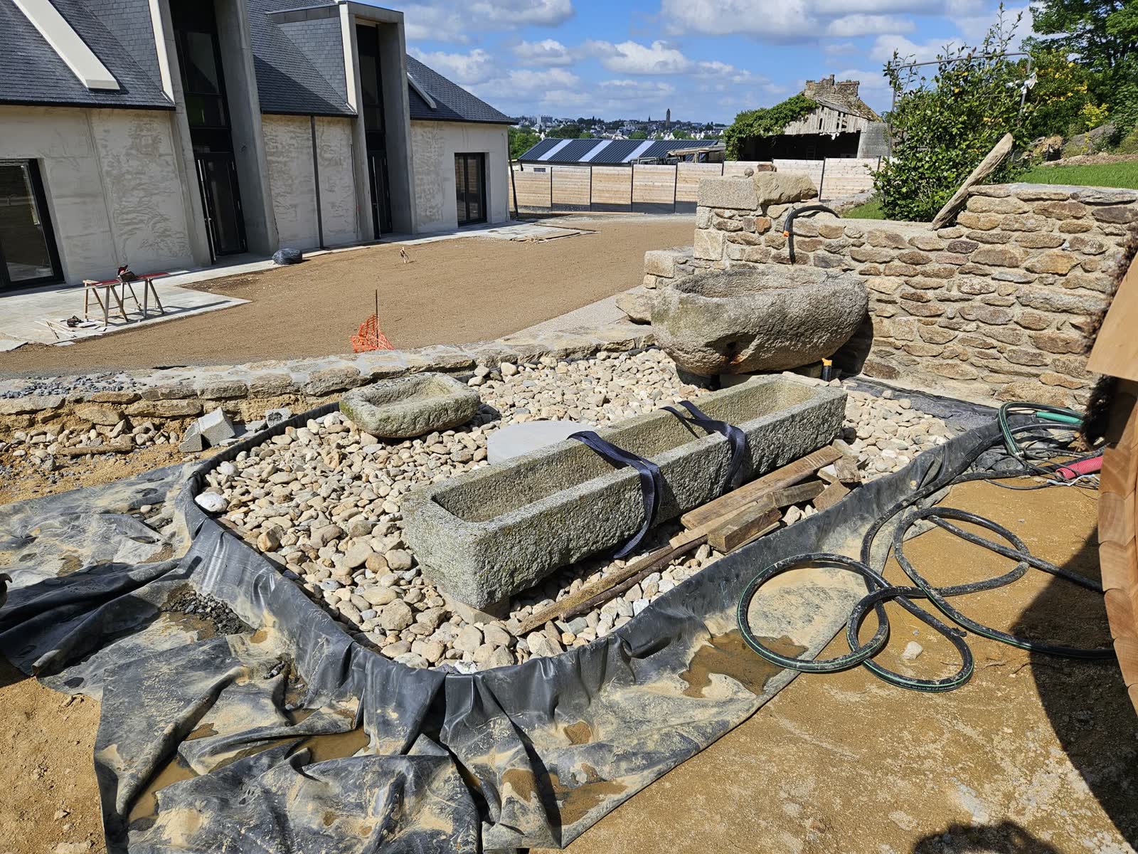 Le chantier en mai 2025: maison neuve, terrasse bois en cours, les auges posées sur un lit de galets, un mur de pierre en construction