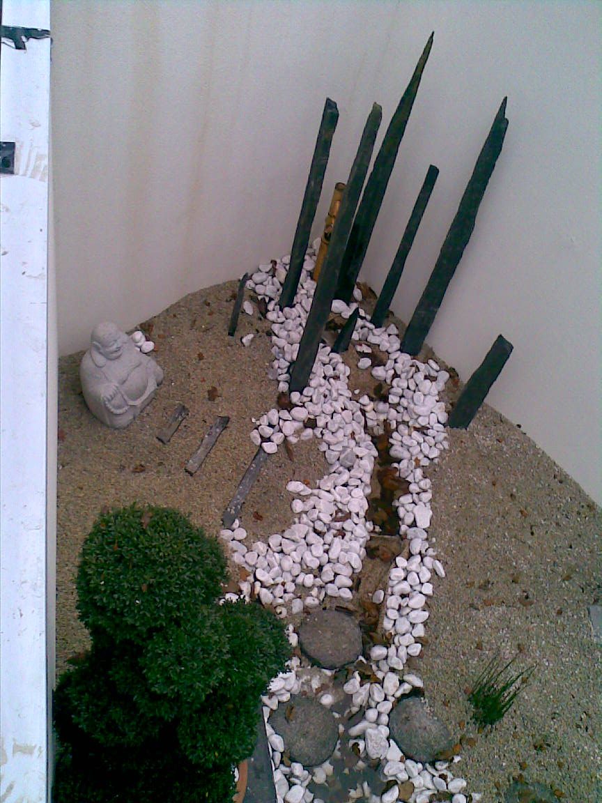 Vue de dessus: les pieux de schiste noir plantés en faisceau, le Bouddha de pierre assis dans le gravier, le ruisseau de galets blancs et une boule de buis