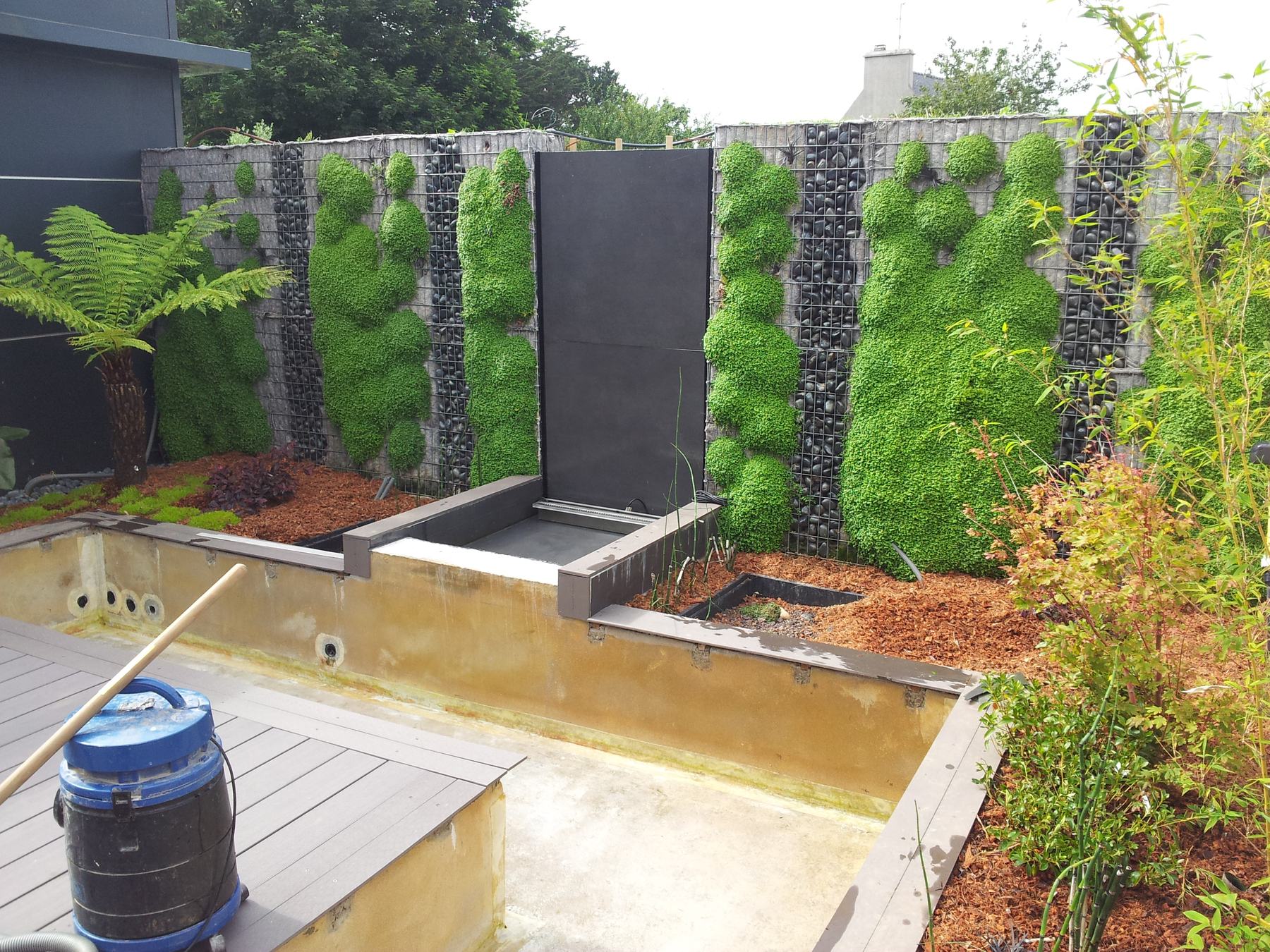 Le même mur en chantier: la maçonnerie brute encore apparente, le bassin non revêtu, la terrasse en cours de pose, un seau de chantier au premier plan