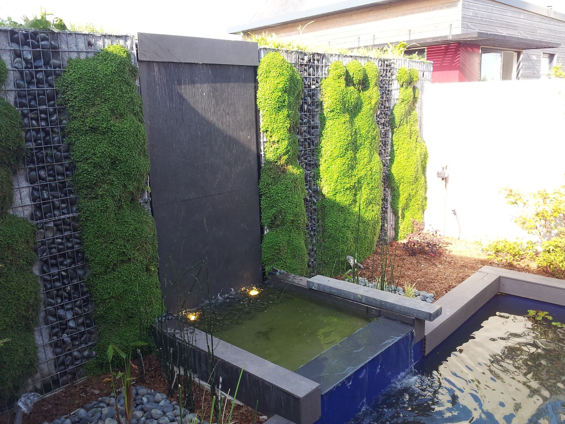 Le mur vu de trois quarts: la dalle d’ardoise sombre où l’eau descend, les colonnes de mousse et les gabions de galets noirs de part et d’autre, la cuve bleue en pied
