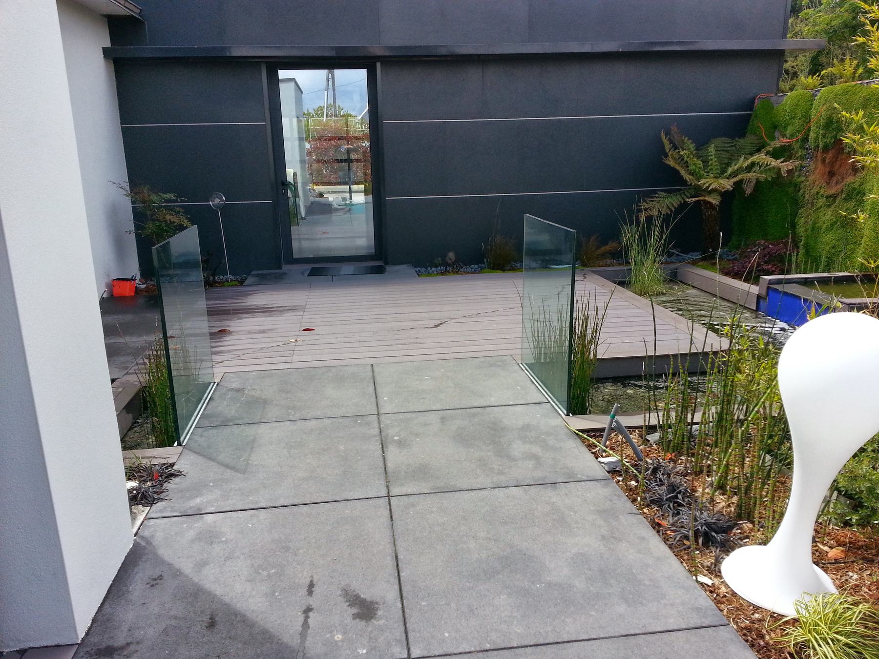 L’entrée de la maison: un dallage d’ardoise franchit le bassin, un garde-corps de verre le borde, la porte vitrée au fond