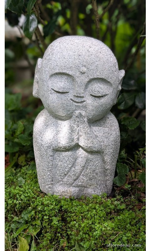 Petit jizo de granit, les mains jointes et les yeux clos, assis dans un tapis de mousse et de feuillage