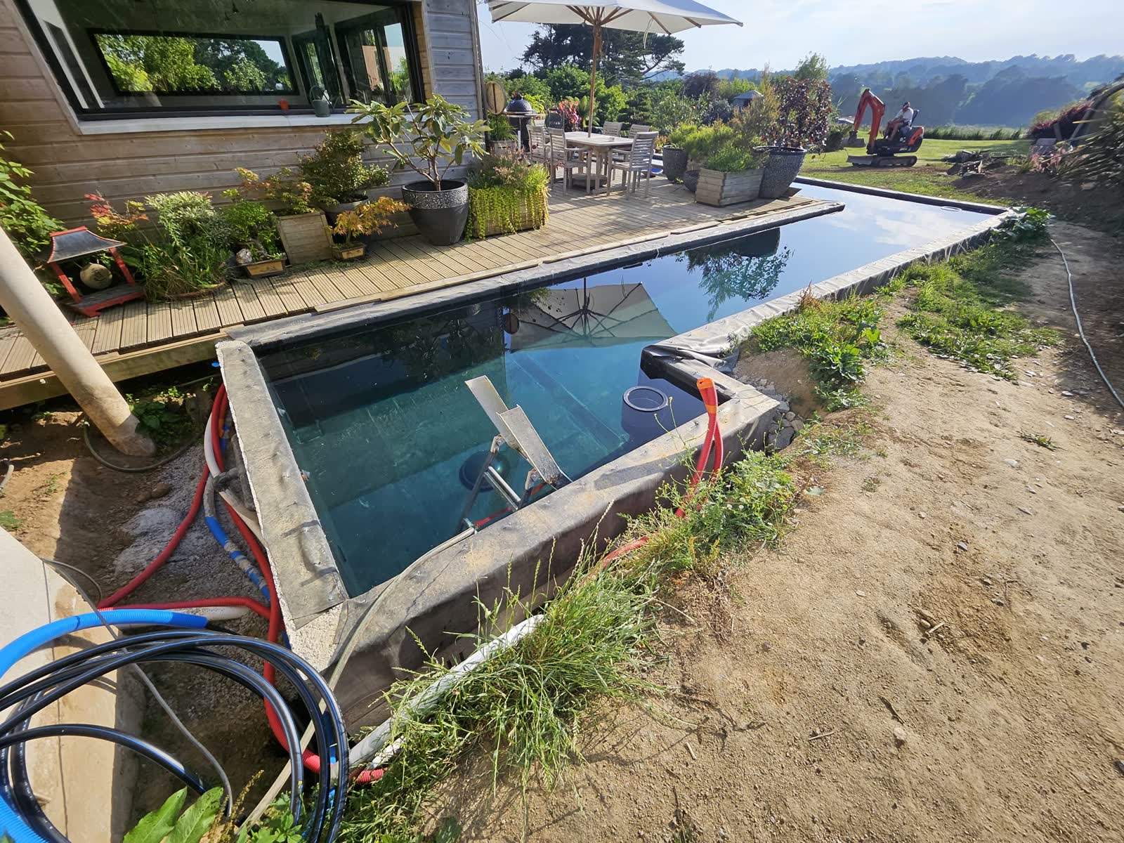 Le bassin en eau vu du haut du terrain: la terrasse bois et ses pots, la maison en bois, la mini-pelle encore sur place, la vallée au fond