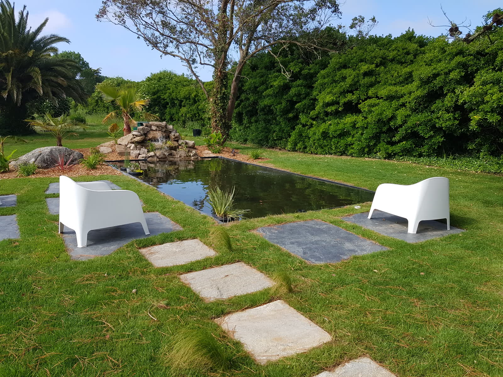 Le bassin fini: un plan d’eau rectangulaire, deux fauteuils blancs de part et d’autre sur des dalles, la cascade de rochers au fond contre la haie, un palmier à gauche