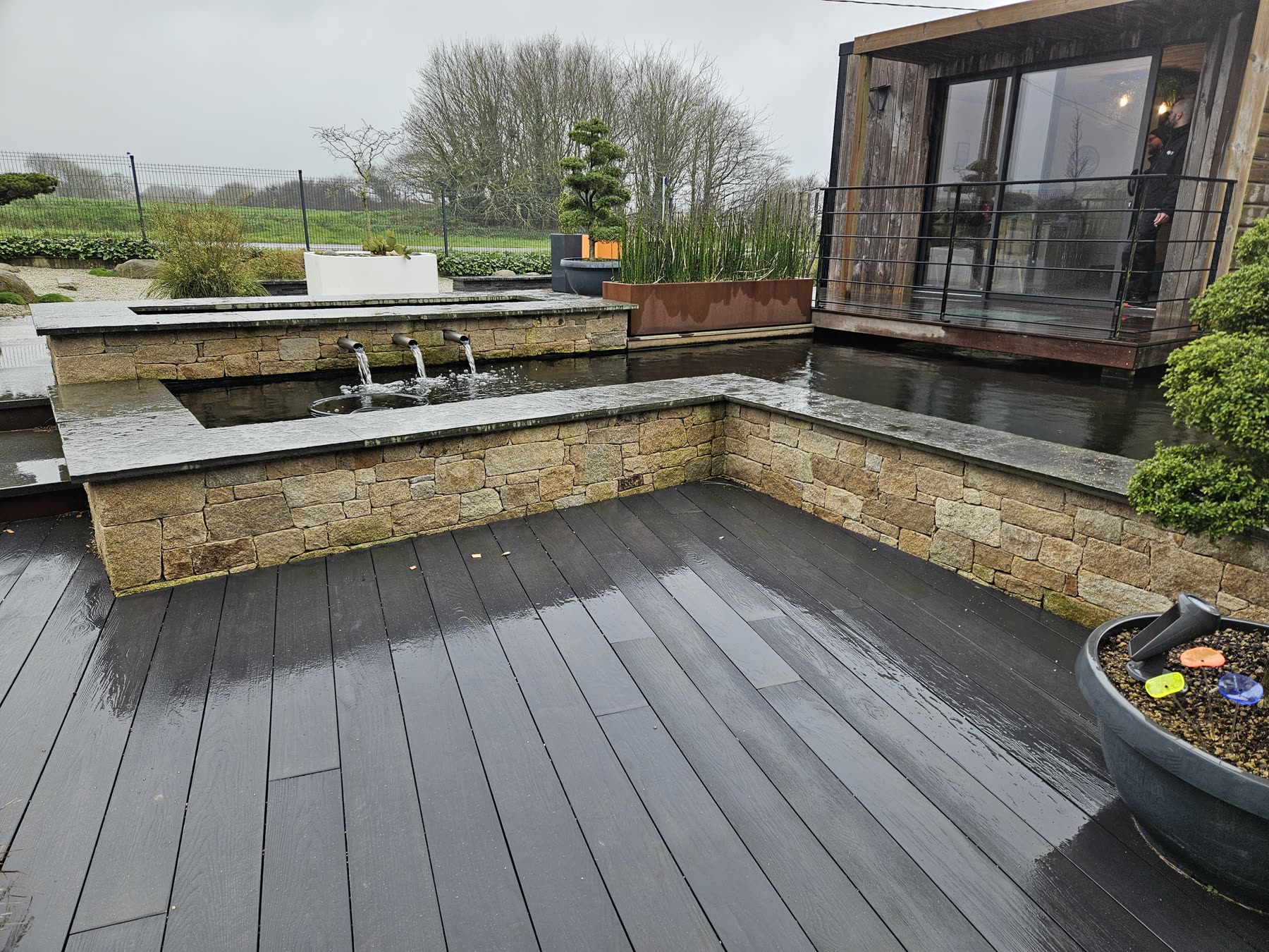 Sous la pluie: une terrasse en lames grises, un mur bas en moellons de granit avec trois becs qui coulent dans un bassin en L, un bureau de jardin en bois avec baie vitrée, des pins taillés en nuage derrière