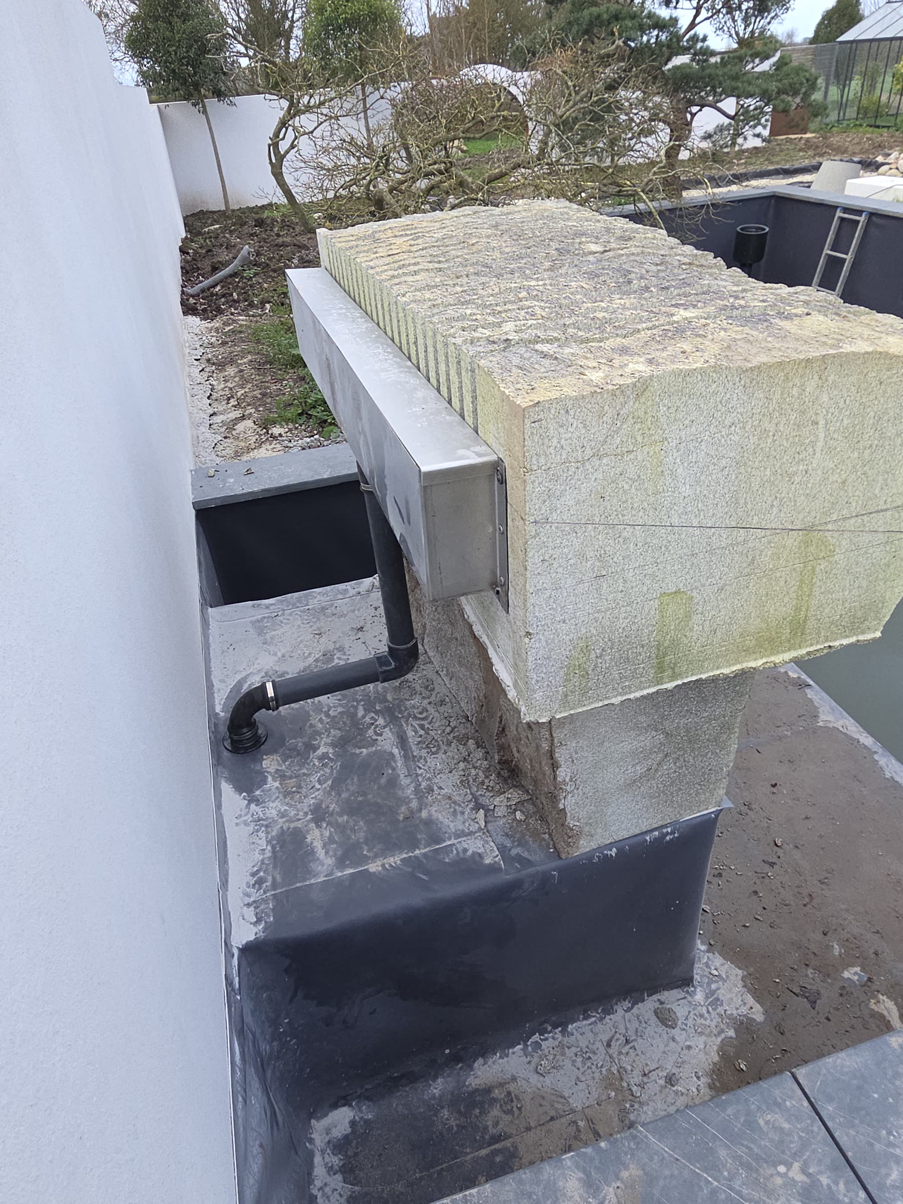Un gros bloc de granit strié posé sur un pilier maçonné, une lame d’eau inox encastrée dessous, la membrane noire du bassin vide, un tuyau PVC noir, un mur blanc à gauche et des pins en nuage au fond