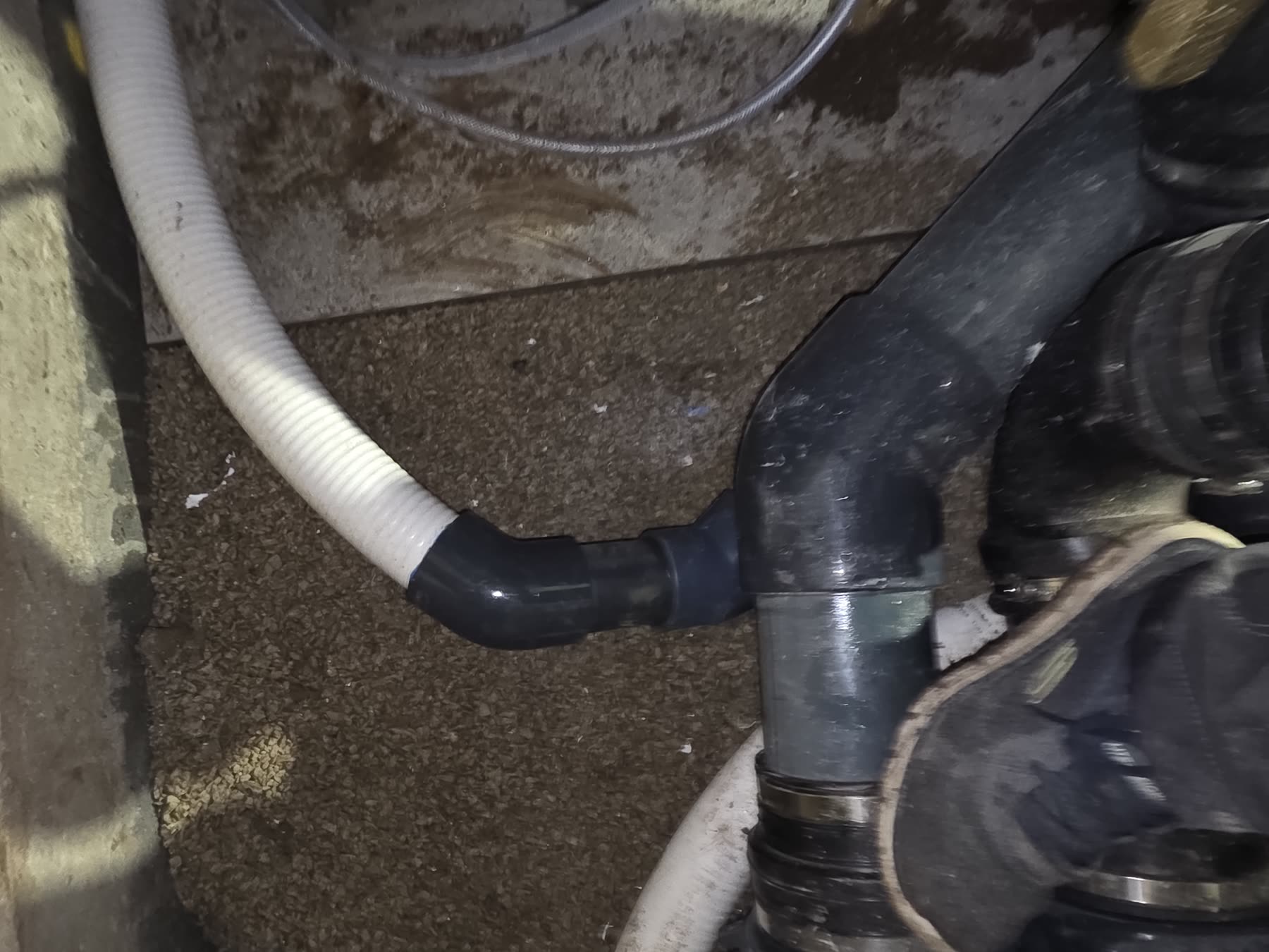 Au sol du local technique: un flexible annelé blanc raccordé par un coude PVC gris à une conduite noire, gravier autour