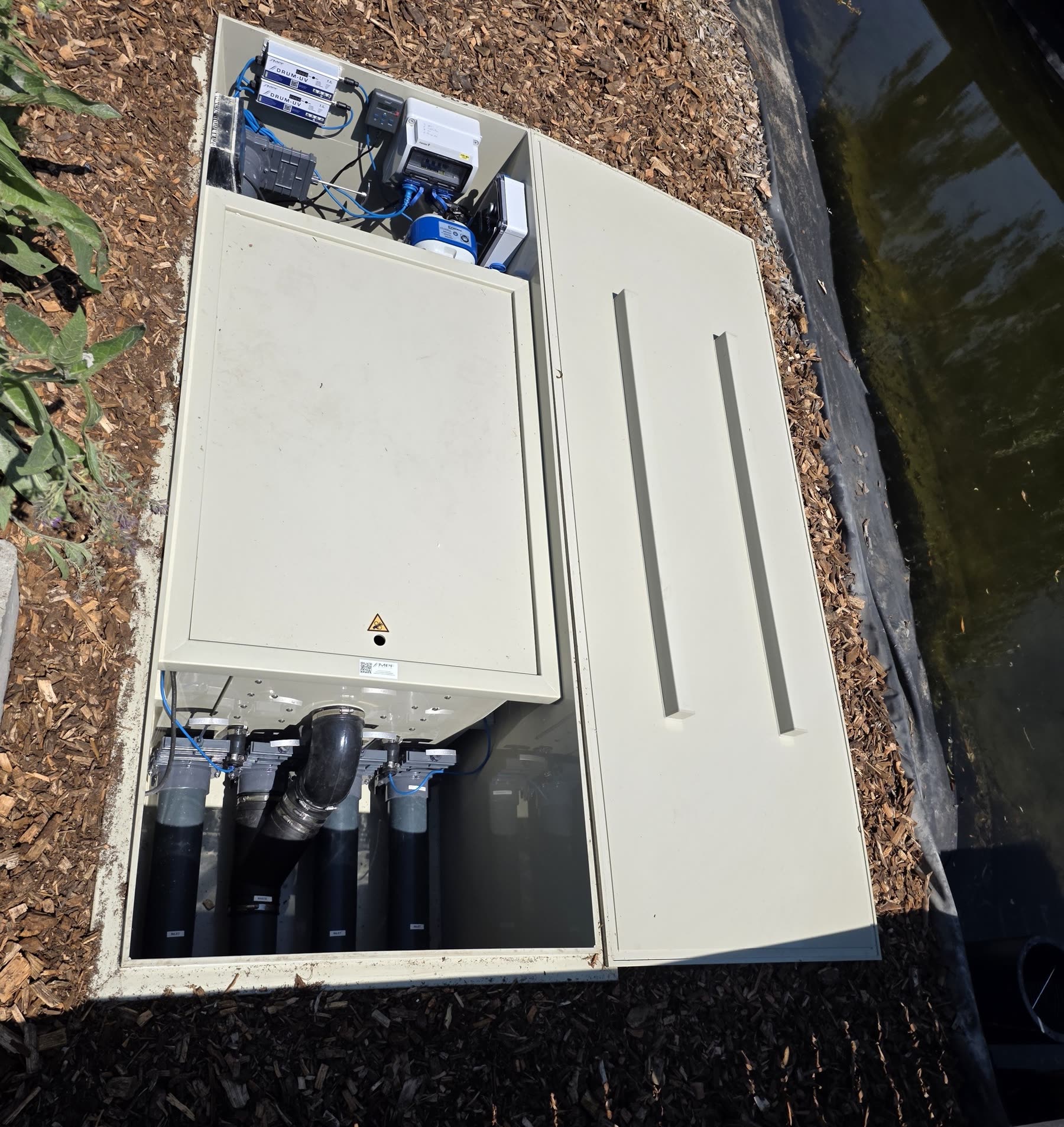 Le local technique enterré, capot ouvert: l’armoire électrique en haut, les canalisations en dessous, paillage de copeaux autour