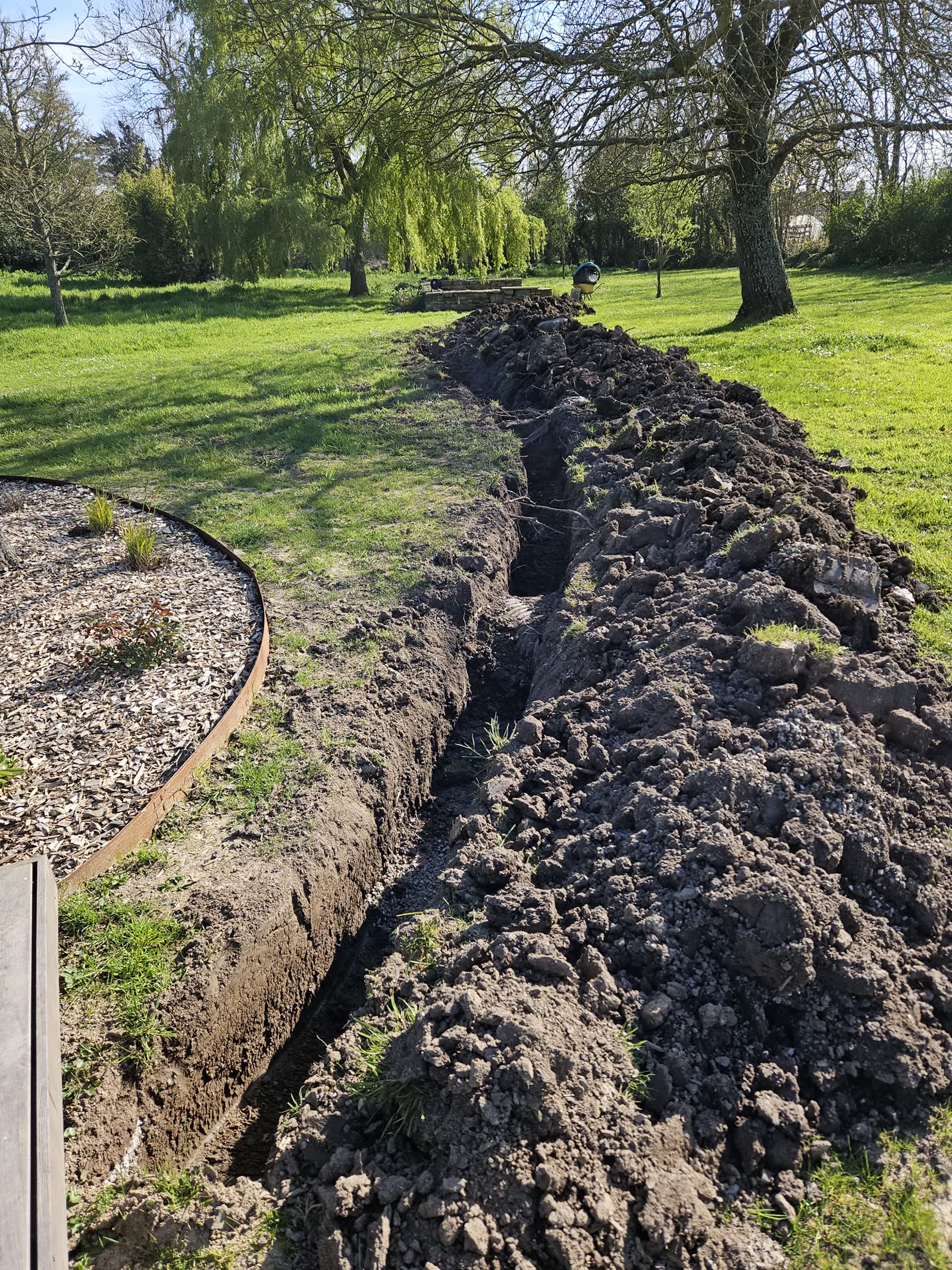 La tranchée d’alimentation ouverte à travers la pelouse, la terre en cordon sur le côté, un massif paillé au premier plan, de grands arbres au fond