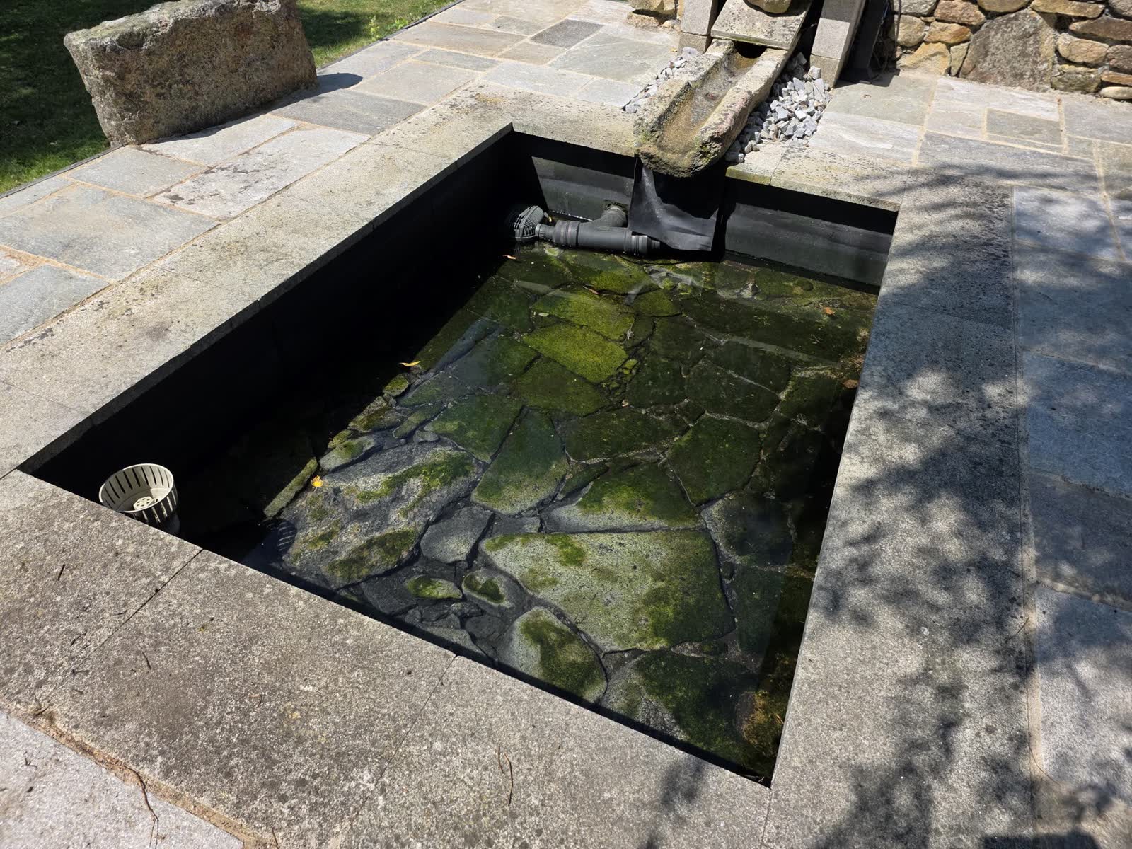 Le lavoir remis en eau: un bassin de pierre en longueur, son dallage de granit autour, l’eau verte sur les dalles du fond, le tuyau d’arrivée d’eau posé dans l’angle