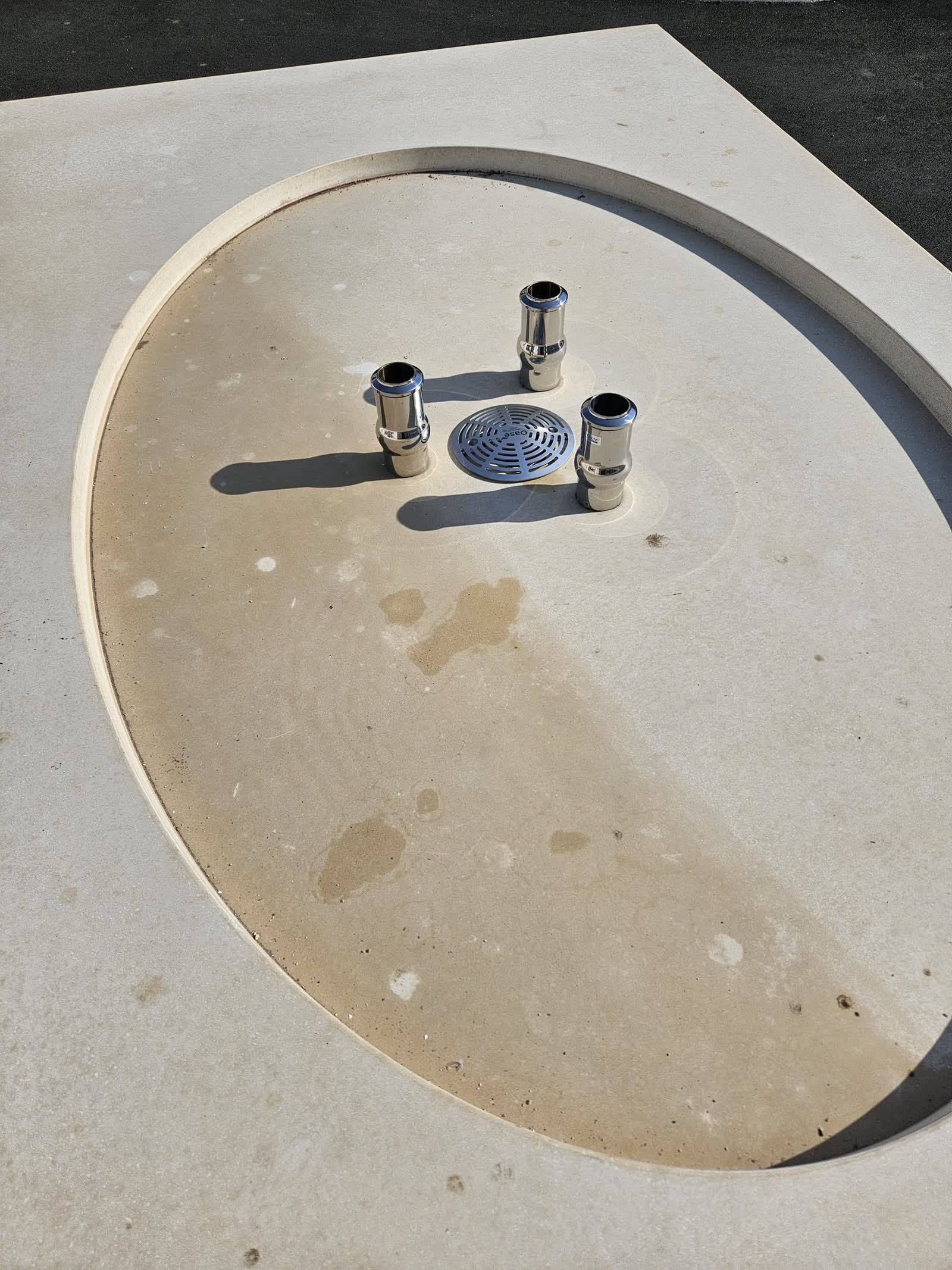 Le bassin ovale à sec, creusé dans la dalle de béton: trois embouts chromés dressés autour d’une bonde ronde grillagée au centre