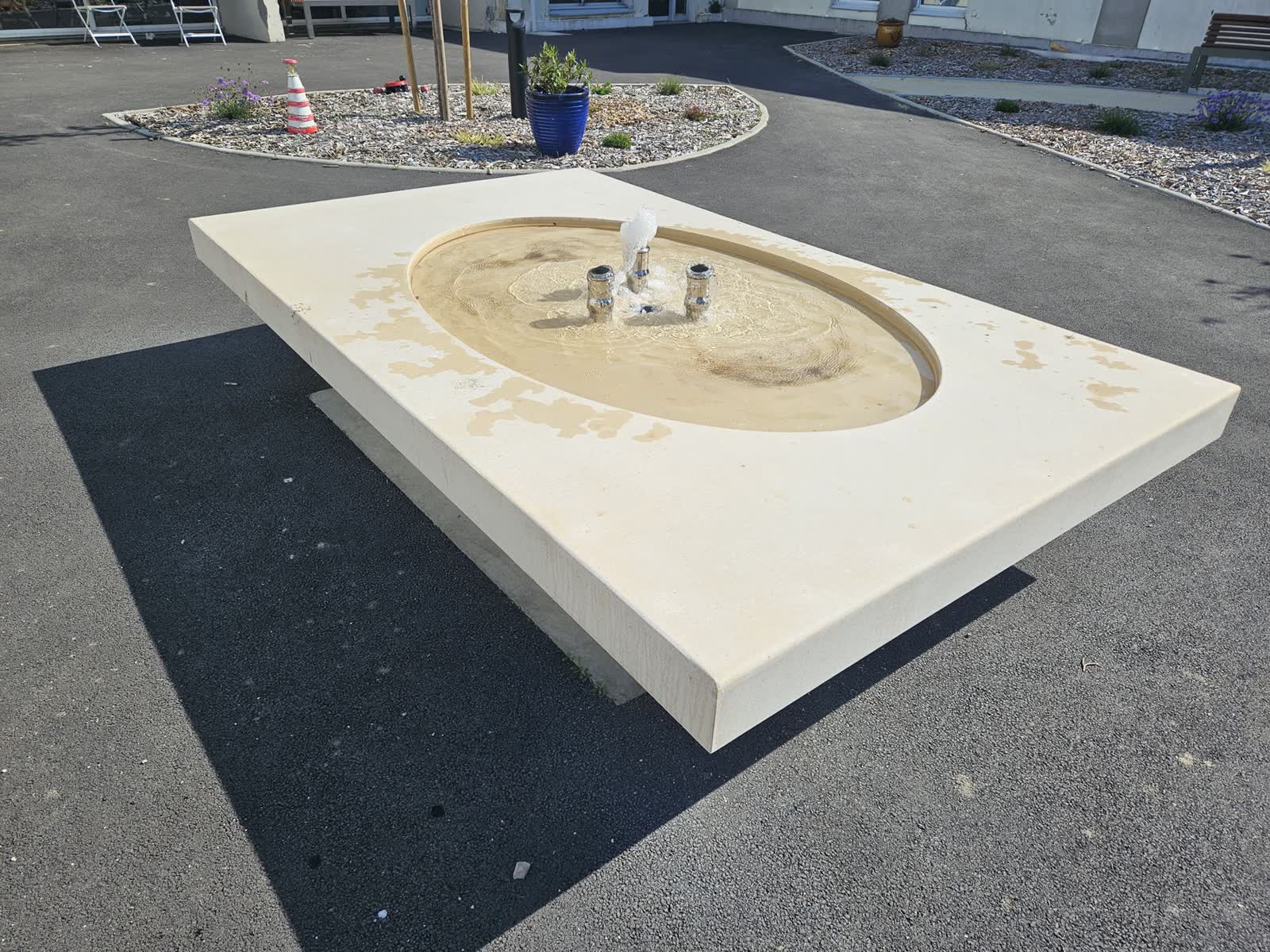 Une dalle en béton clair posée sur un socle dans une cour en enrobé, un bassin ovale creusé dedans, les trois jets en marche, des massifs de graviers et un bâtiment blanc derrière