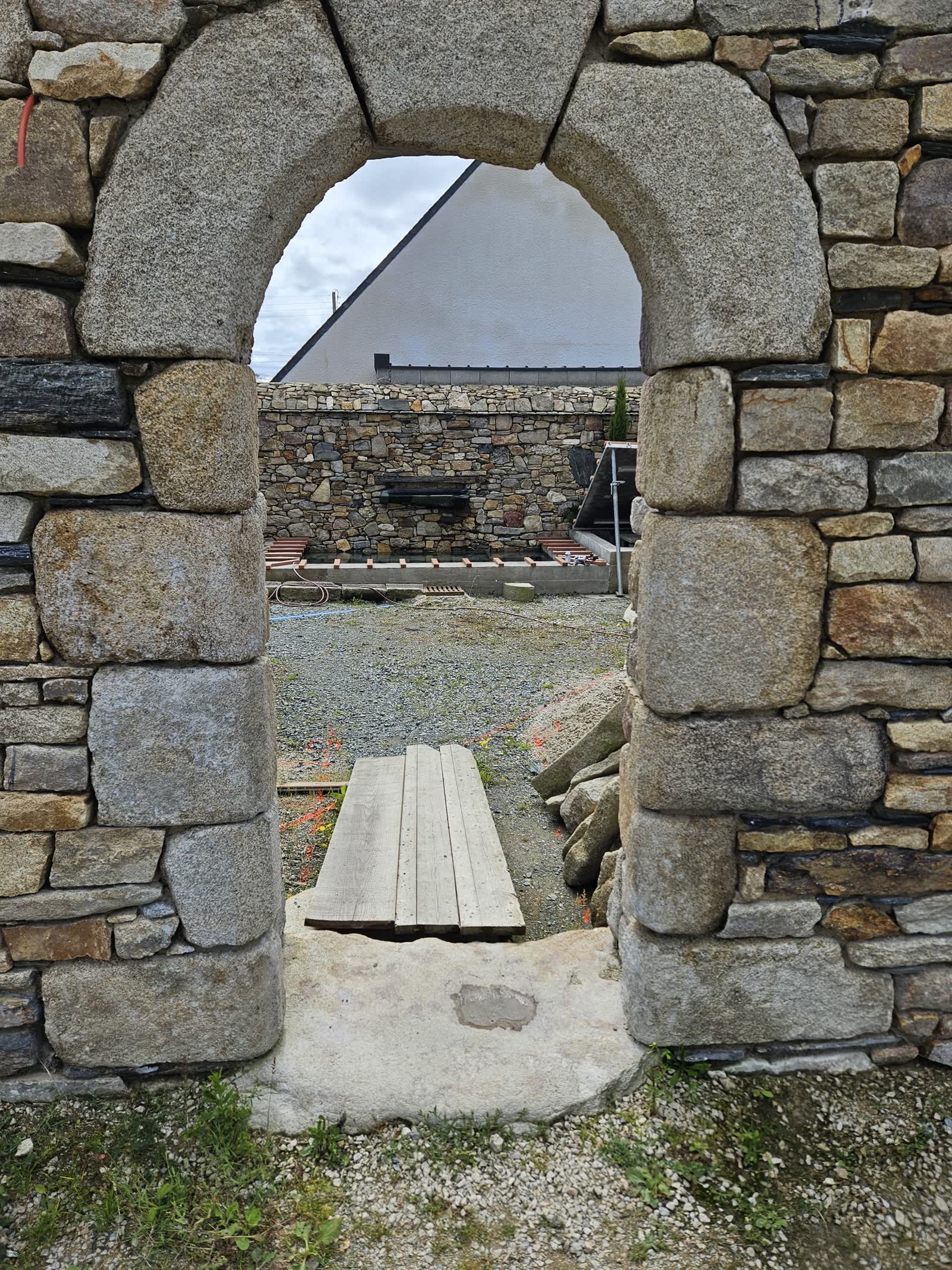 Une arche en granit dans un mur en pierre sèche ouvre sur le patio en chantier: gravier, planches au sol, le mur du fond avec l’ouverture de la lame d’eau, un pignon de maison derrière