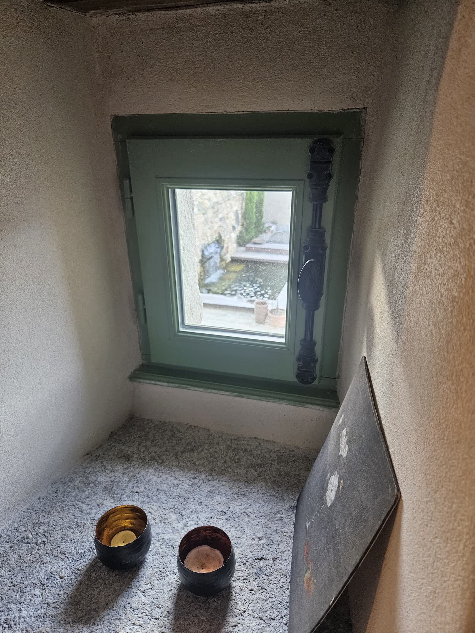 Une petite fenêtre à cadre vert dans un mur enduit, deux photophores sur l’appui en granit; par la vitre, on voit le bassin du patio et le mur de pierre